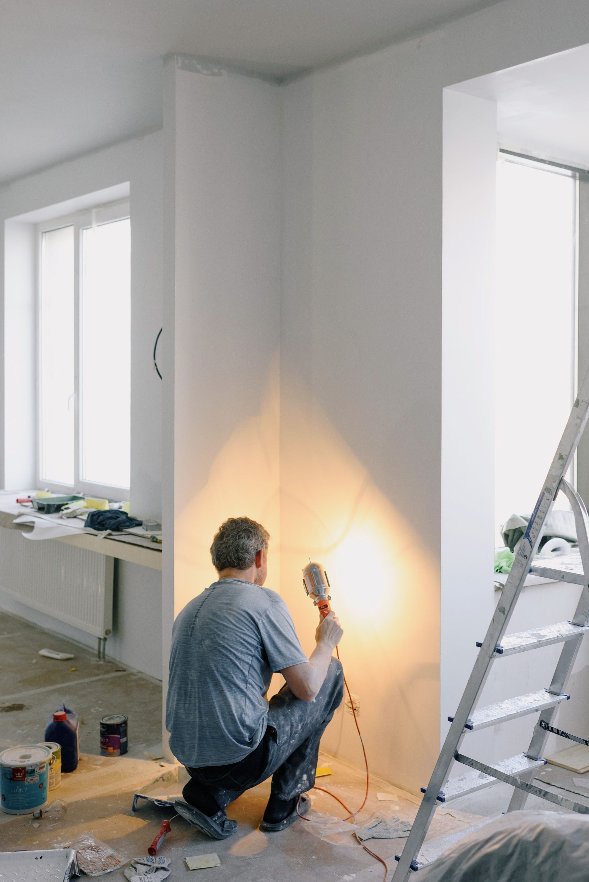Tradesperson kneeling to finish a wall corner with a heat tool in a freshly painted room, with a ladder and paint supplies nearby