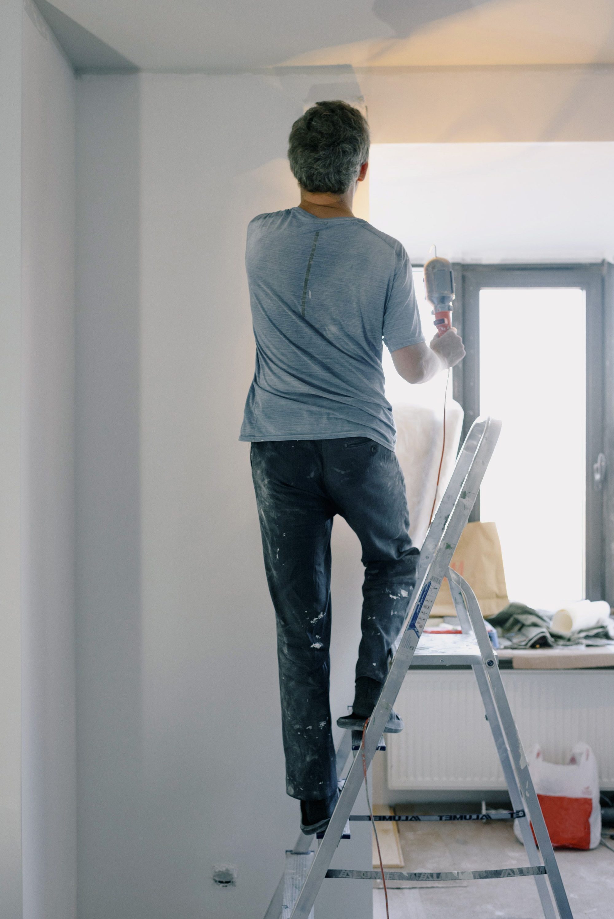 Worker on a stepladder reaching up to finish a ceiling corner, paint-marked work pants and a hand tool in use