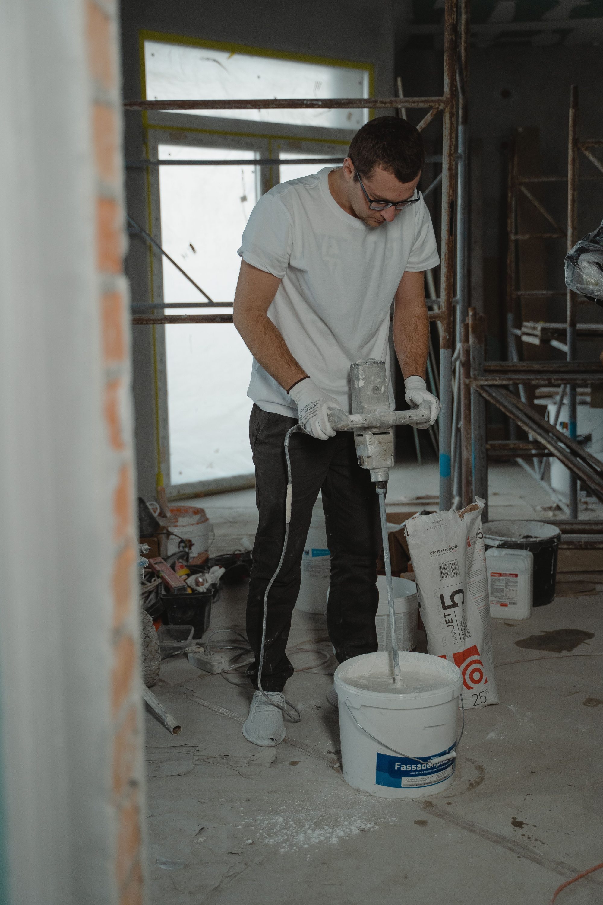 Contractor mixing plaster compound with a paddle mixer beside scaffolding and a brick wall opening at a commercial job site