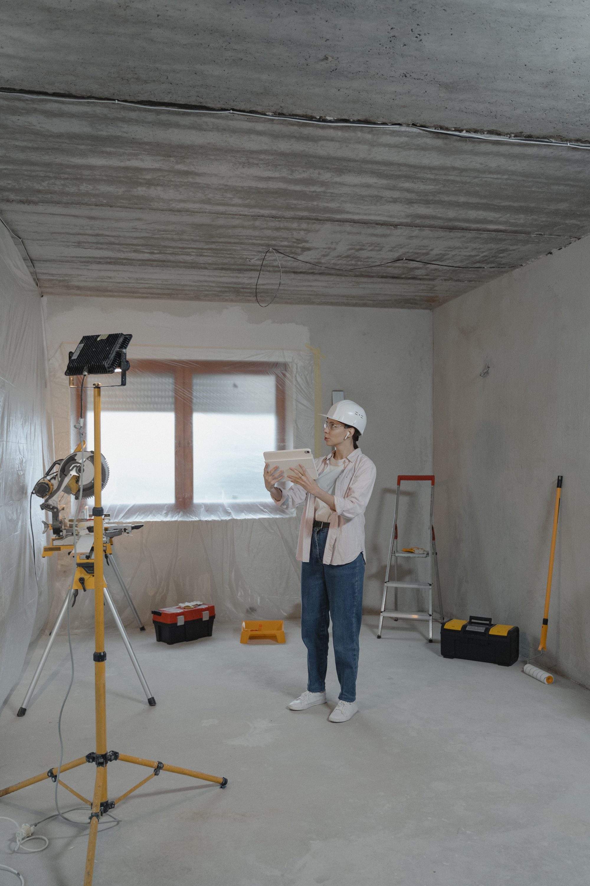 Site supervisor in a hard hat and safety glasses reviewing project plans on a tablet in an unfinished concrete room, with a miter saw station and tool boxes nearby